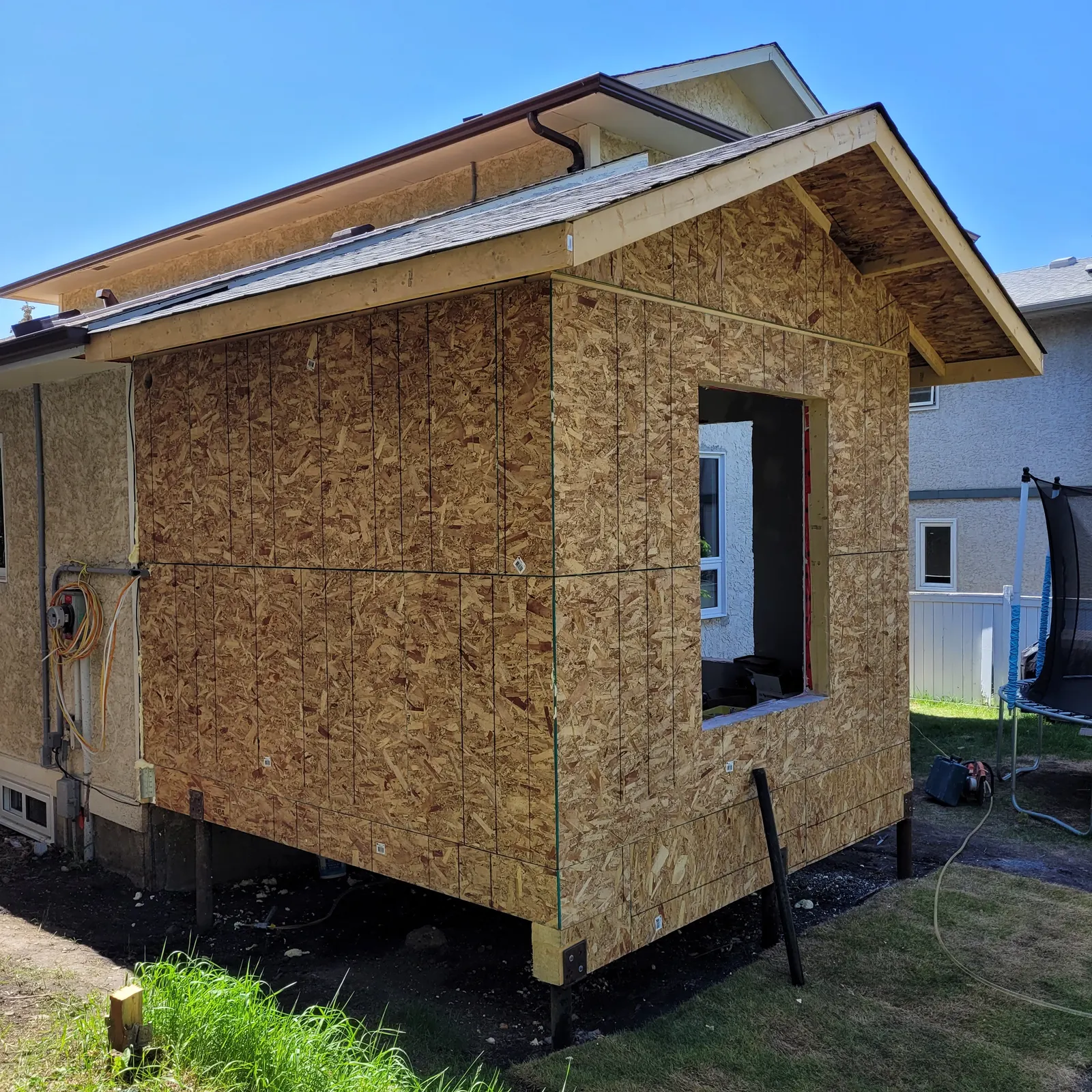 Rear house addition during construction with exposed wood sheathing, roof framing and an unfinished window opening beside the existing house.