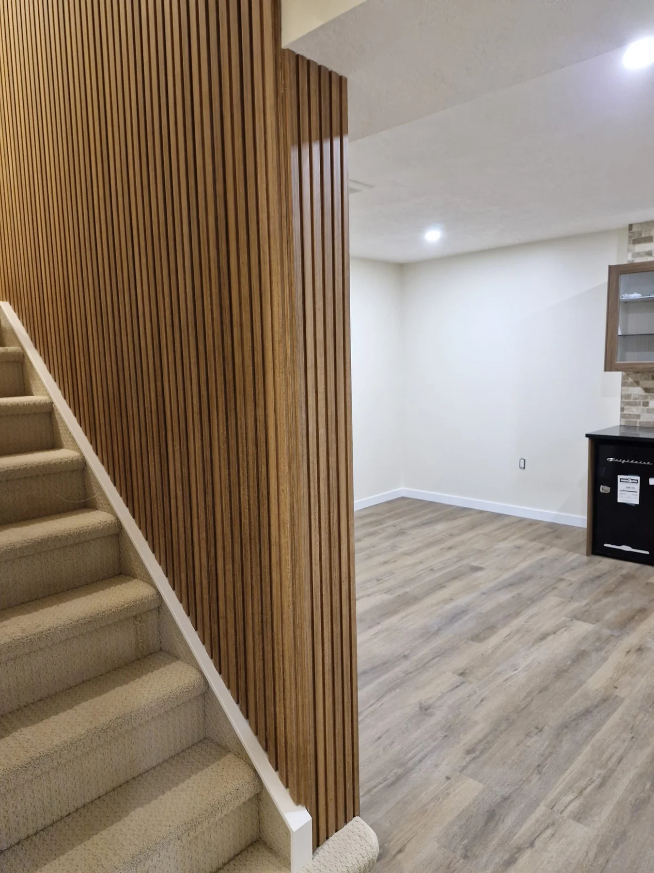 Renovated basement with carpeted stairs, a vertical wood-slat feature wall, wood-look flooring and a wet bar.