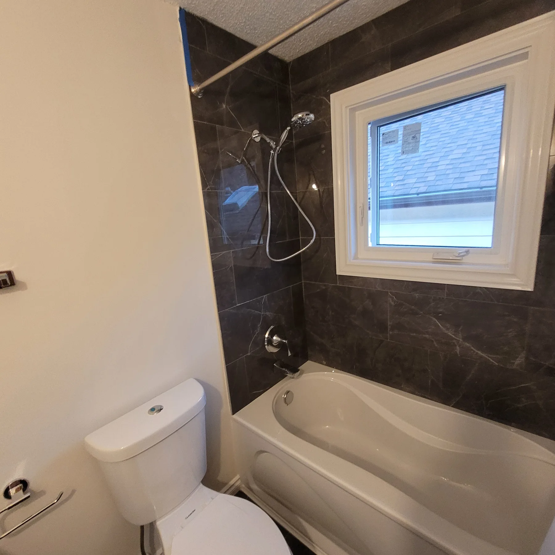 White bathtub with dark veined tile surround, chrome handheld shower and white-framed window.