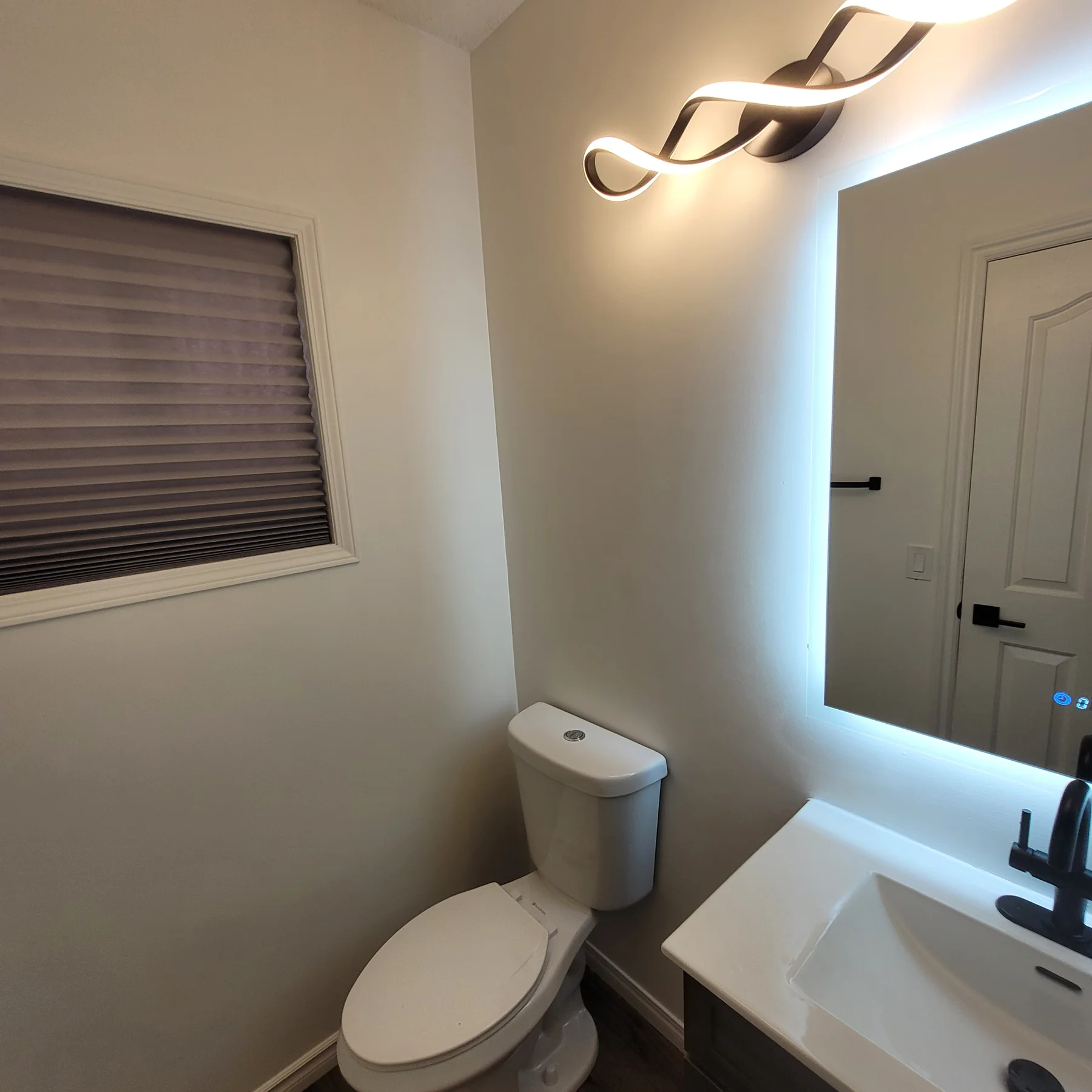 Finished bathroom with white toilet, window blind, neutral walls and an illuminated mirror beside the vanity.