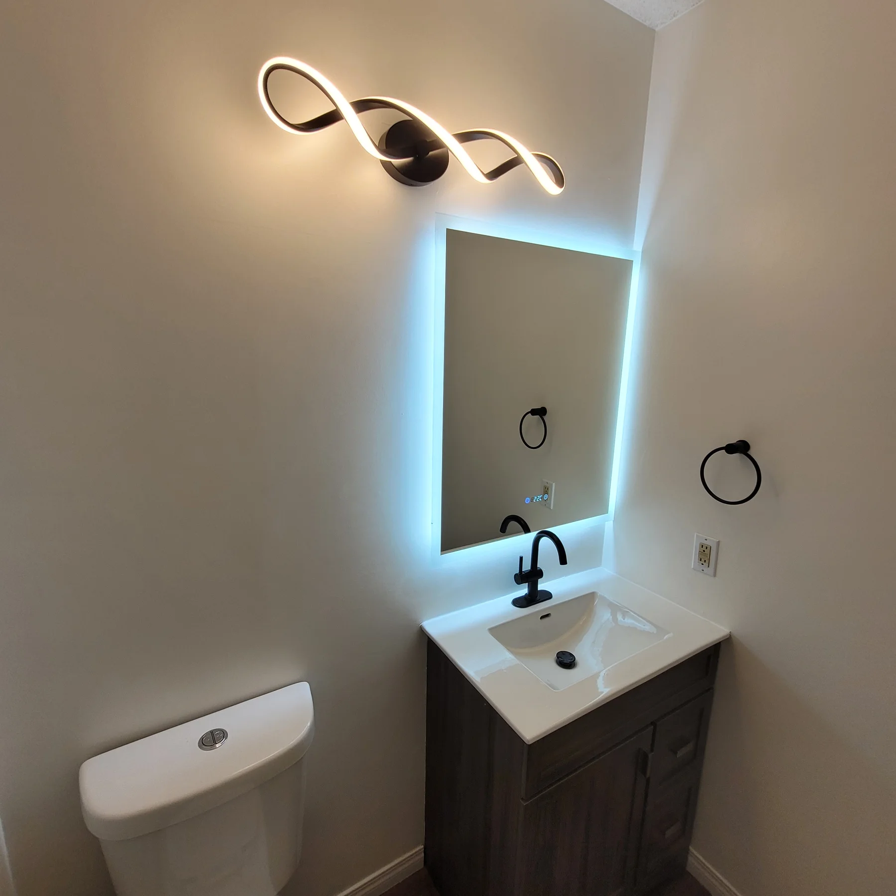Renovated bathroom vanity with dark cabinetry, a white sink, black faucet, backlit mirror and curved wall light.