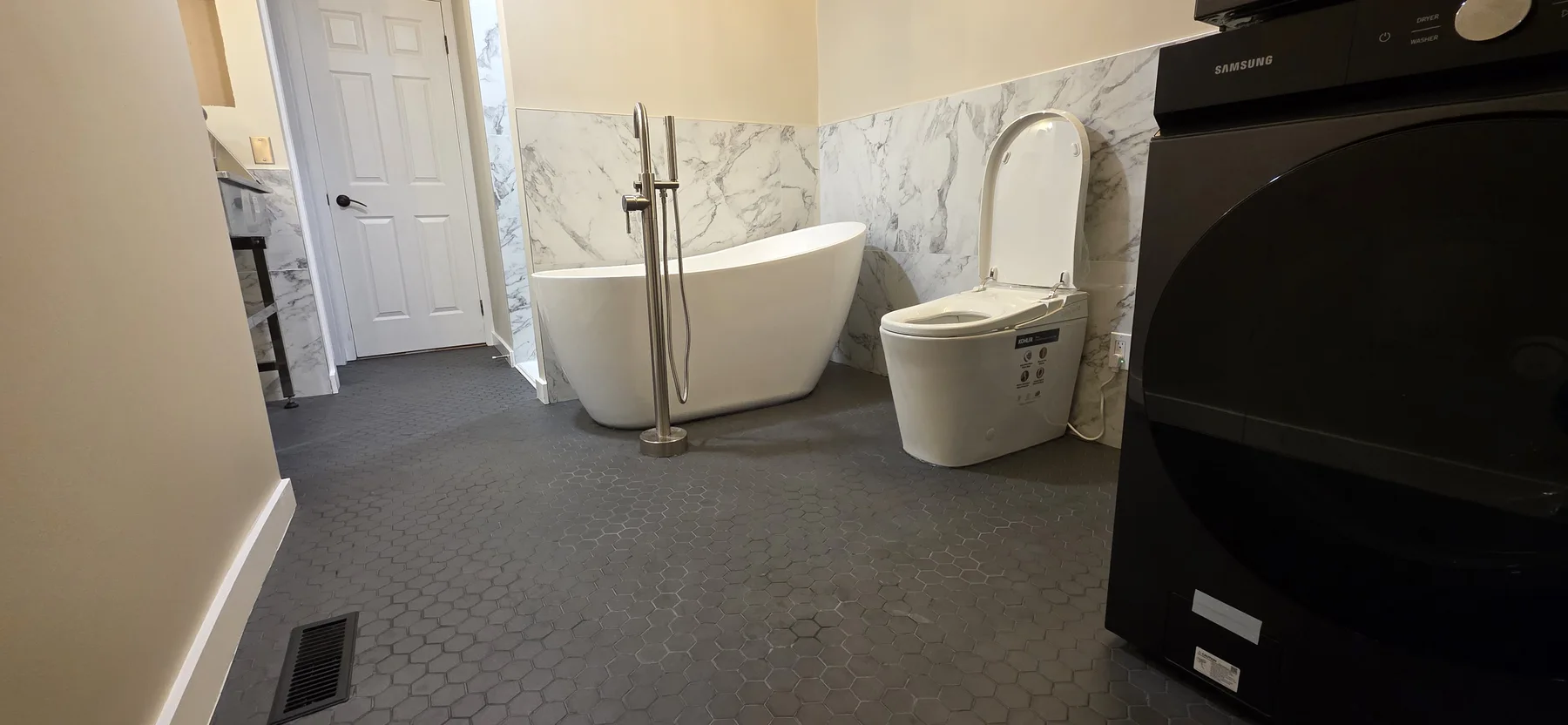 White freestanding bathtub with floor-mounted faucet, marble-look wall tile and dark hexagonal floor beside stacked laundry appliances.