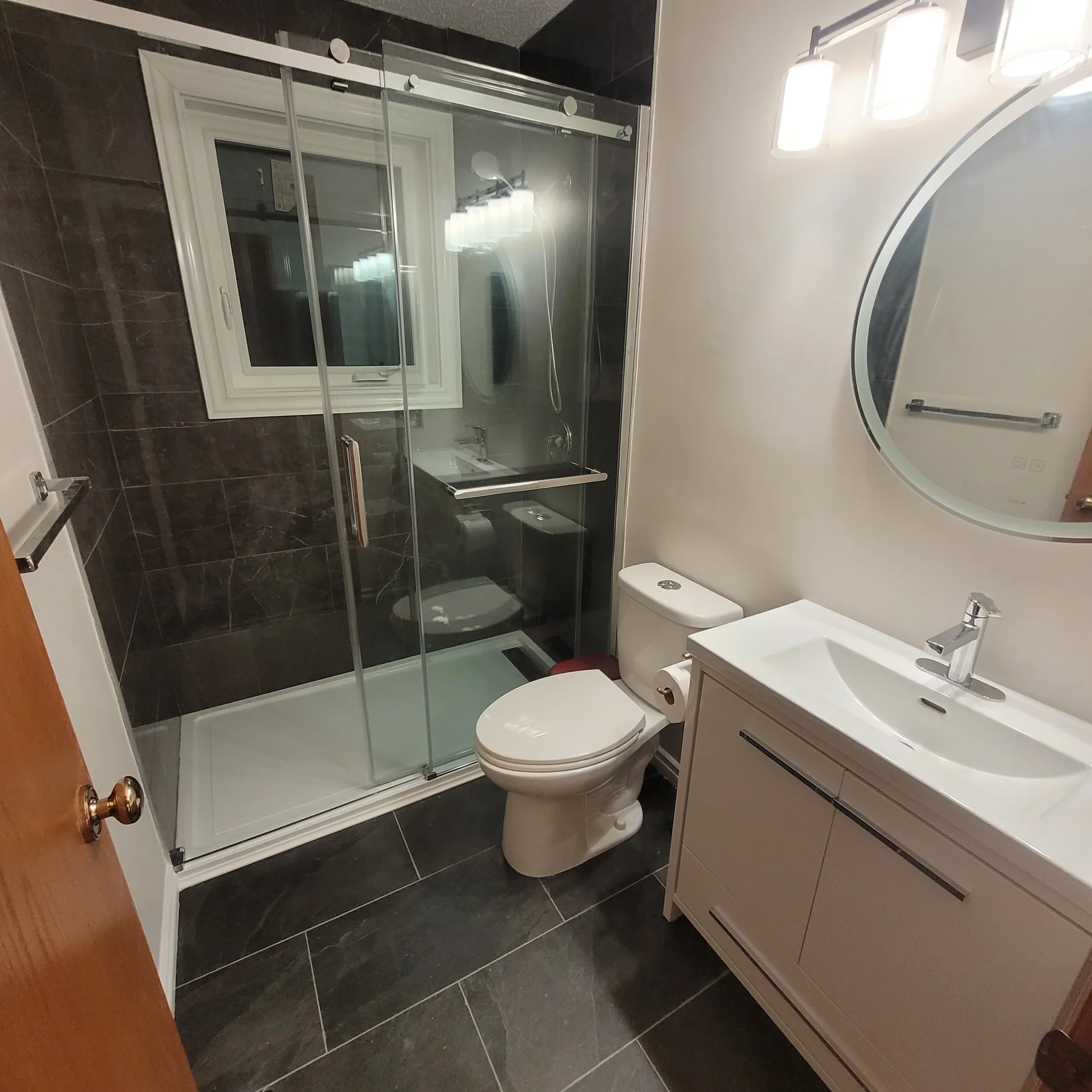 Renovated bathroom with sliding glass shower doors, dark tile, grey vanity, white sink and round mirror.