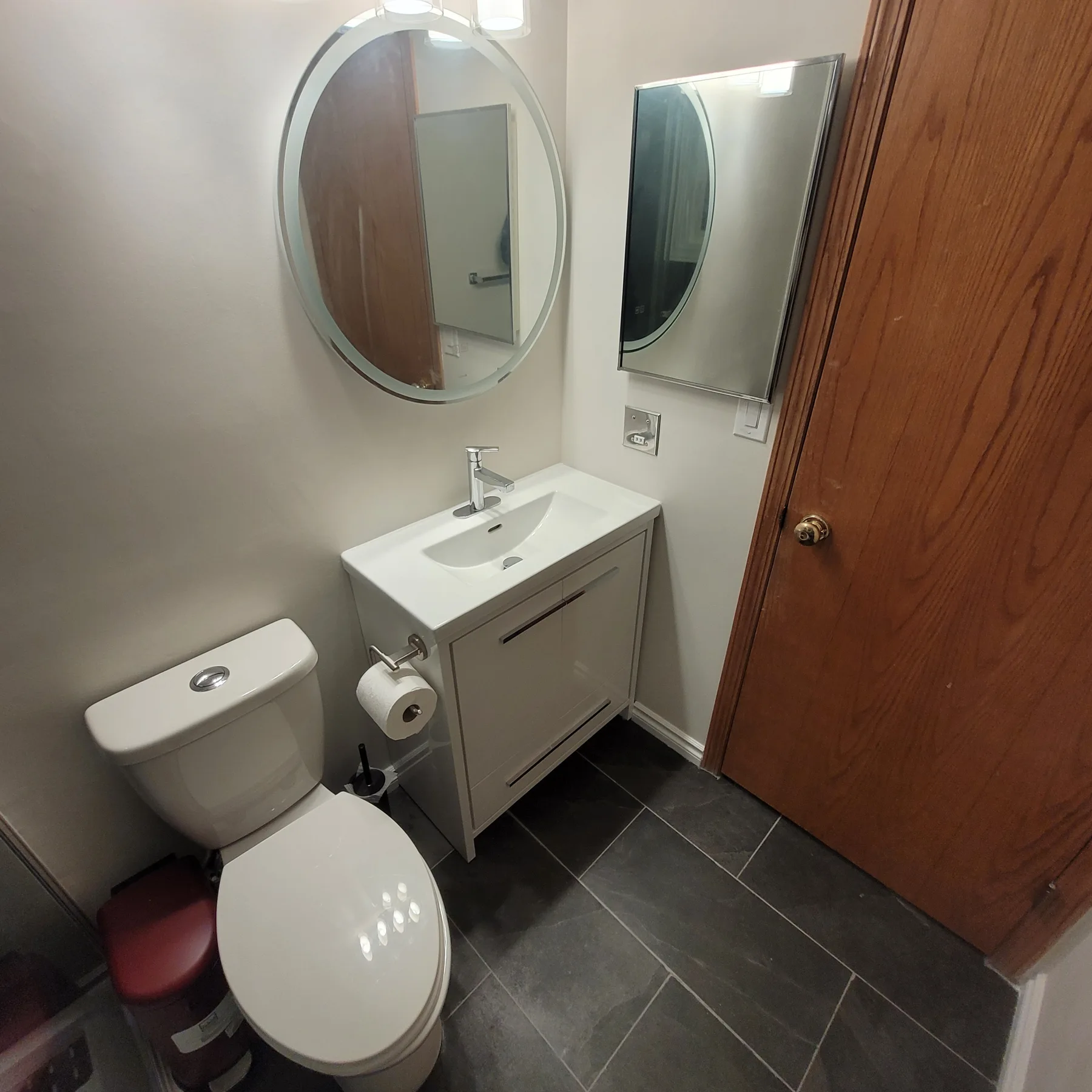 Light grey bathroom vanity with white basin, chrome faucet, round mirror and dark tiled floor.