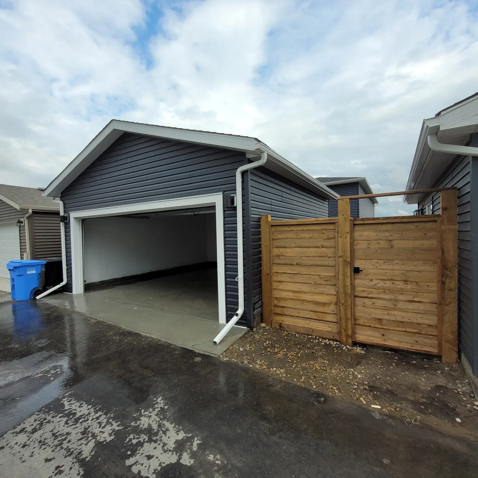 Detached garage with blue-grey siding, white gable trim and gutters, an open overhead door and an adjoining wood privacy fence.