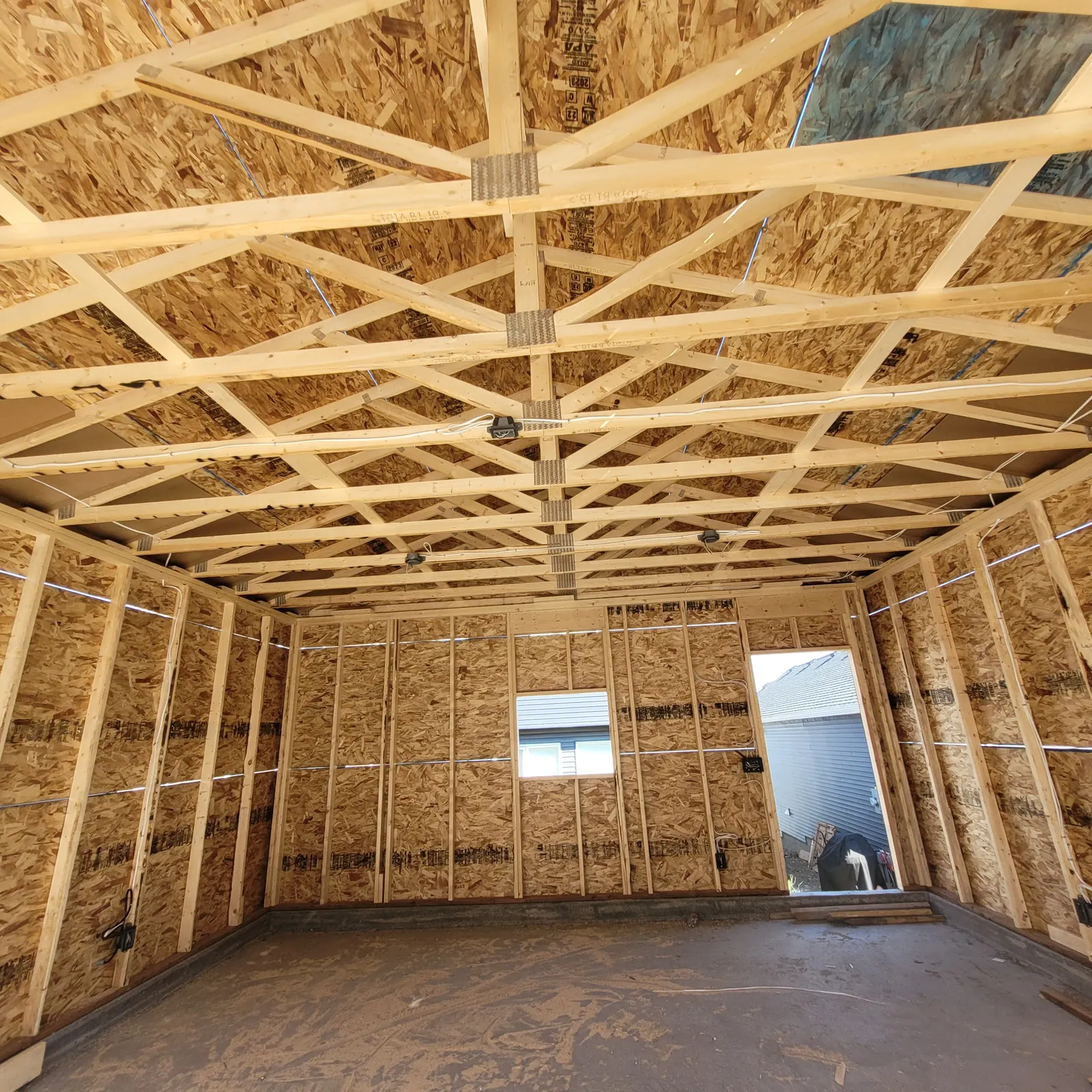 Detached garage interior under construction with exposed wood wall studs, roof trusses, sheathing, a window opening and an entry-door opening.