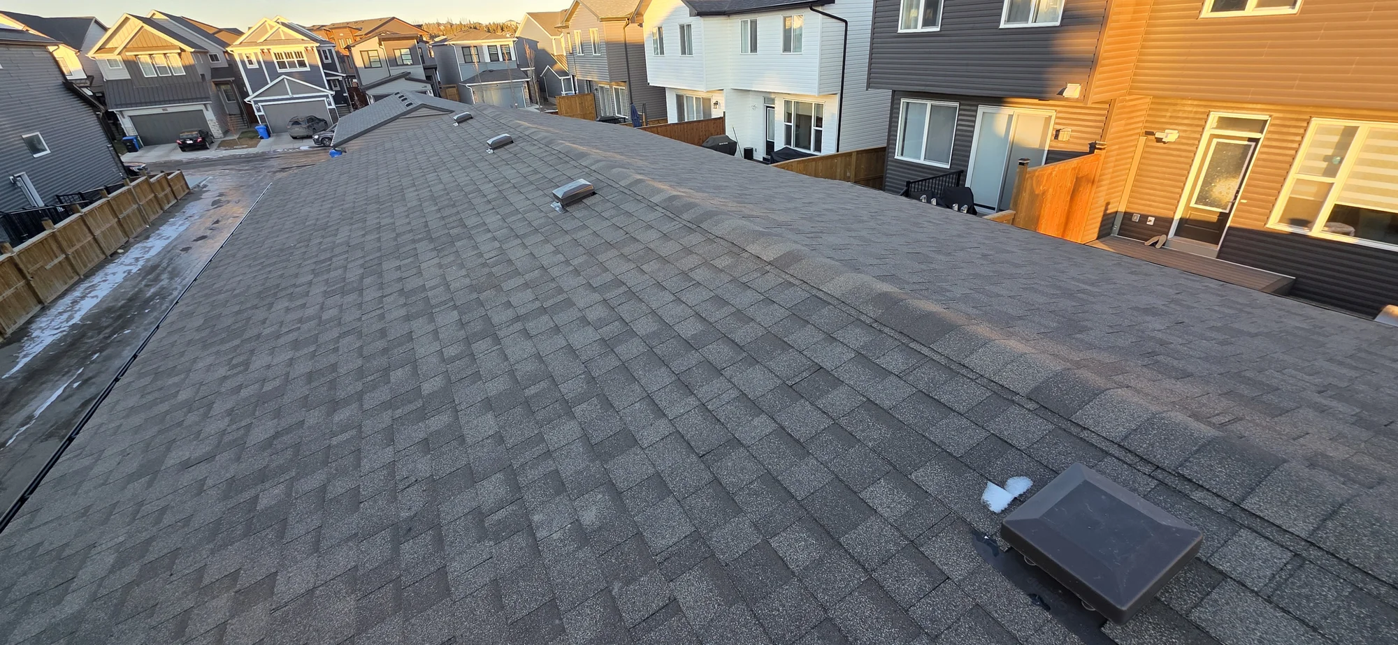 Garage roof with grey asphalt shingles, ridge caps and roof vents, viewed from above with neighbouring homes in the background.