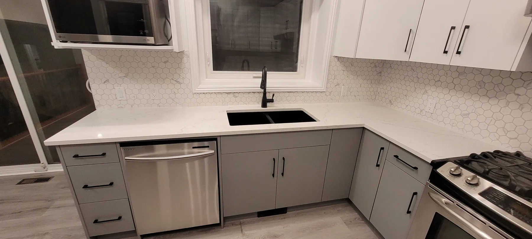 Renovated kitchen sink area with grey lower cabinets, white upper cabinets, a light countertop, hexagonal-tile backsplash, black sink and faucet, and stainless dishwasher.
