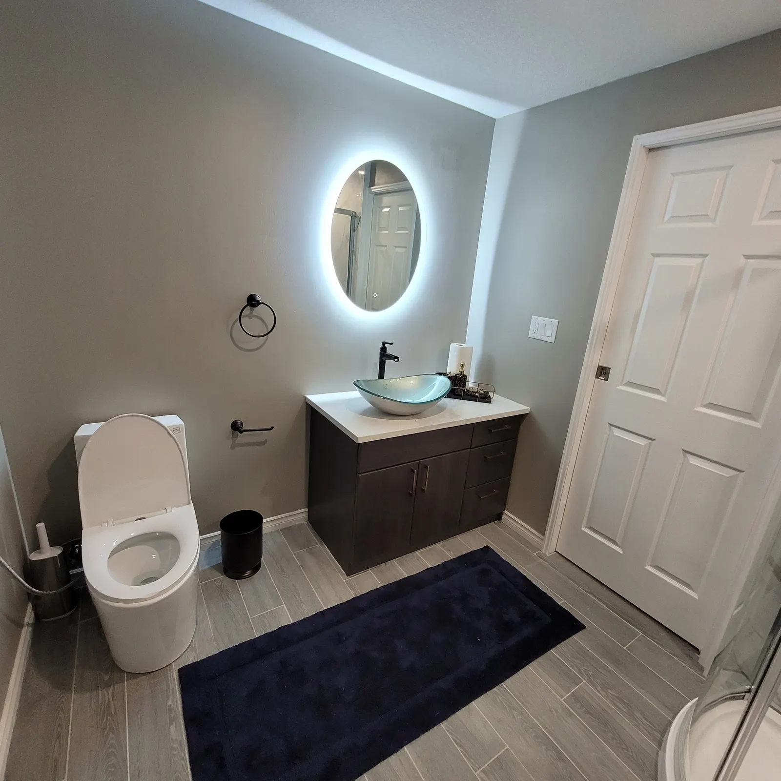 Basement bathroom with a dark floating vanity, glass vessel sink, illuminated oval mirror, toilet, grey floor tiles and a curved glass shower enclosure.