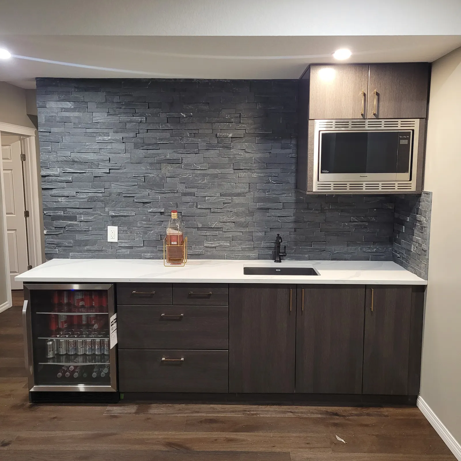 Basement wet bar with dark wood cabinets, a light countertop, textured grey backsplash, black sink and faucet, beverage fridge and stainless microwave.