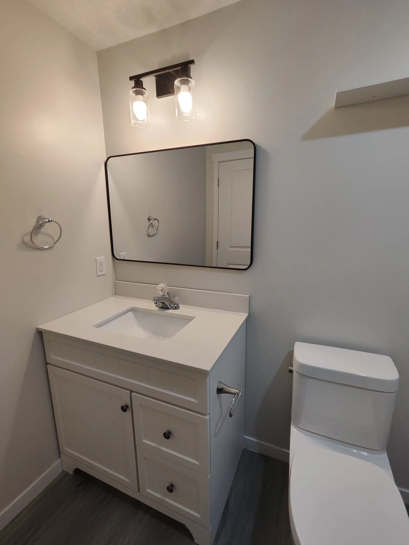 Secondary suite bathroom with a light vanity, white basin, black-framed mirror, two wall lights, white toilet and wood-look flooring.
