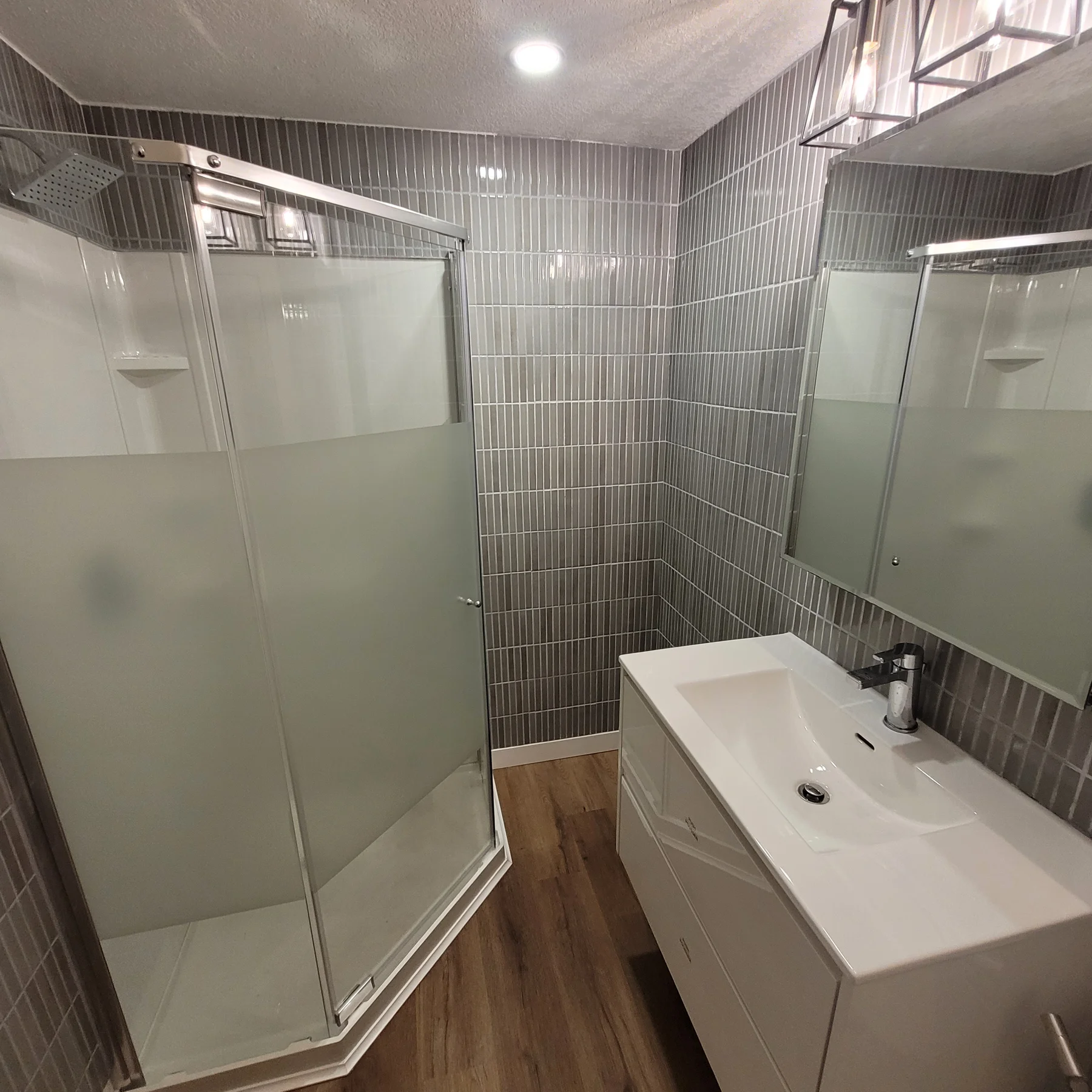 Secondary suite bathroom with a frosted-glass corner shower, vertical grey tile, white vanity, large mirror and wood-look flooring.