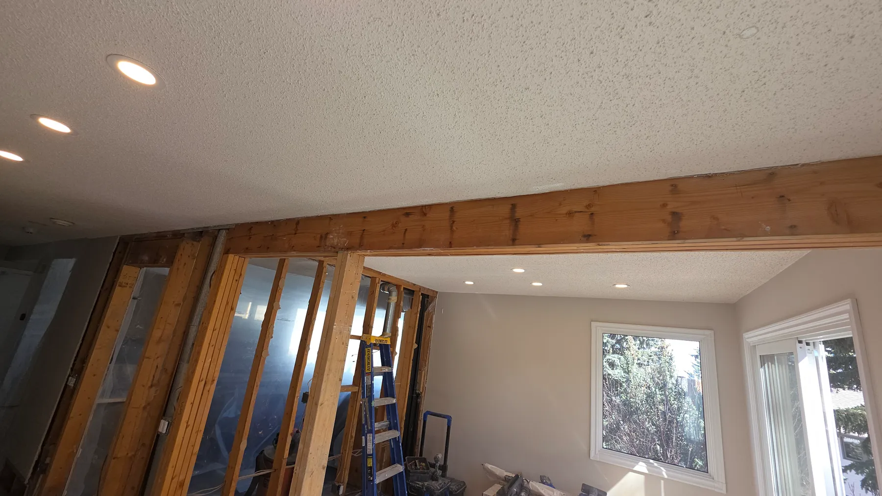 Exposed wood beam and wall framing during the Vincete load-bearing wall removal project, with a ladder and window beyond the opening.