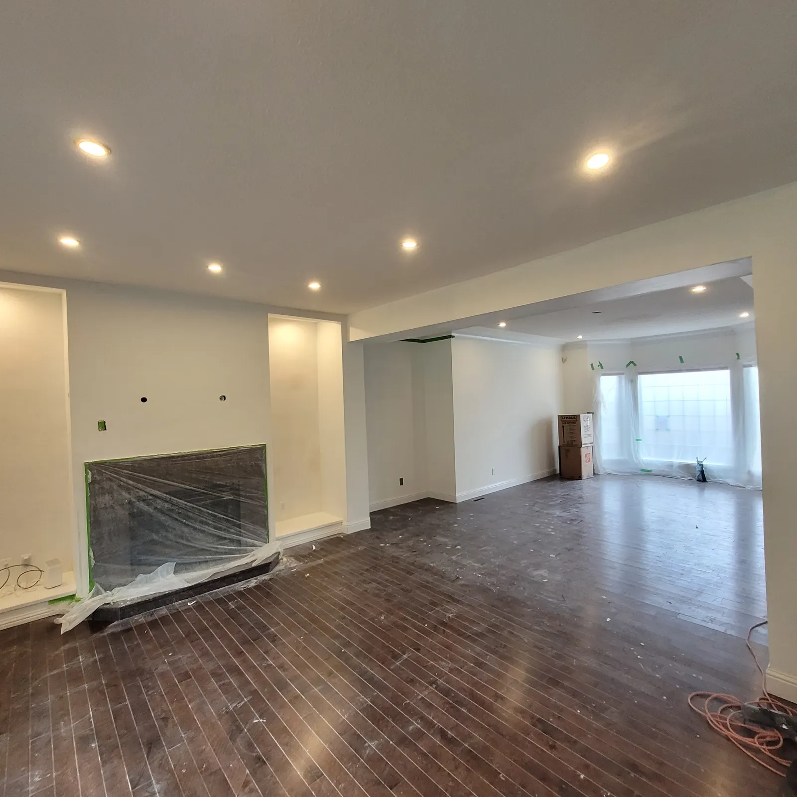 Living room during finishing with an enclosed beam over a wide opening, recessed lights, a protected fireplace and protective film on the wood floor.