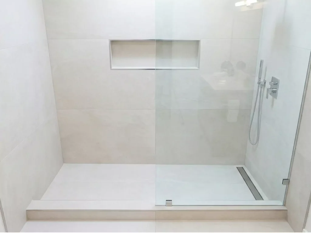 A photo of a modern, spacious walk-in shower featuring large-format beige tiles, a frameless glass enclosure, a built-in shower niche, and a linear drain, making the bathroom feel airy and bright