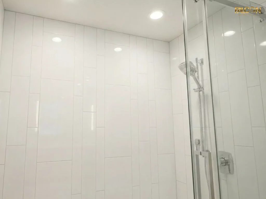 A close-up detail of the shower area showing glossy white subway tiles stacked vertically, reflecting the light from the recessed LED pot lights and emphasizing the ceiling height.