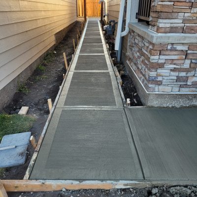 High-angle view of a newly poured concrete walkway running between two houses, featuring a brushed finish and wooden forms, situated next to a wall with multi-colored stone veneer skirting.