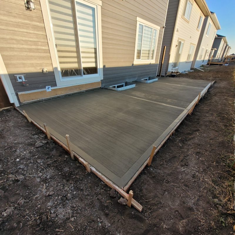 High-angle view of a newly poured concrete patio slab in a backyard, featuring a brushed finish and control joints, surrounded by wooden forms and situated behind a townhouse with beige siding.