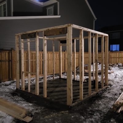 Nighttime view of a backyard construction site showing the wooden framing skeleton of a small detached structure, with snow on the ground and a wooden privacy fence in the background.