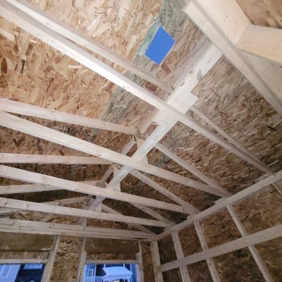 Low-angle interior view of a roof structure under construction, showing a network of wooden trusses supporting OSB sheathing, with a blue vent baffle visible near the ridge.