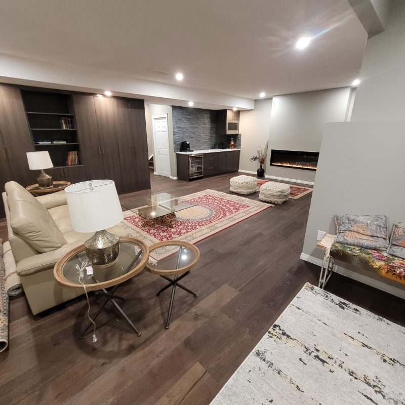 Modern finished basement living area featuring dark hardwood floors, a cream leather sofa, a red traditional area rug, a wet bar with stone backsplash, and a linear electric fireplace.