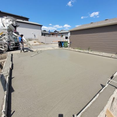 Construction scene showing a concrete mixer truck pouring fresh cement into a framed driveway area, with a worker managing the chute under a bright blue sky.