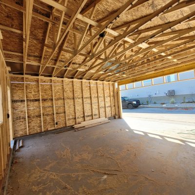 Interior view of a residential garage under construction featuring exposed wooden roof trusses, stud framing with OSB sheathing, and a dusty concrete floor.