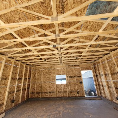 Interior view of a garage framing stage showing engineered wood roof trusses, OSB sheathing on walls with a small window cutout, and electrical wiring rough-ins.