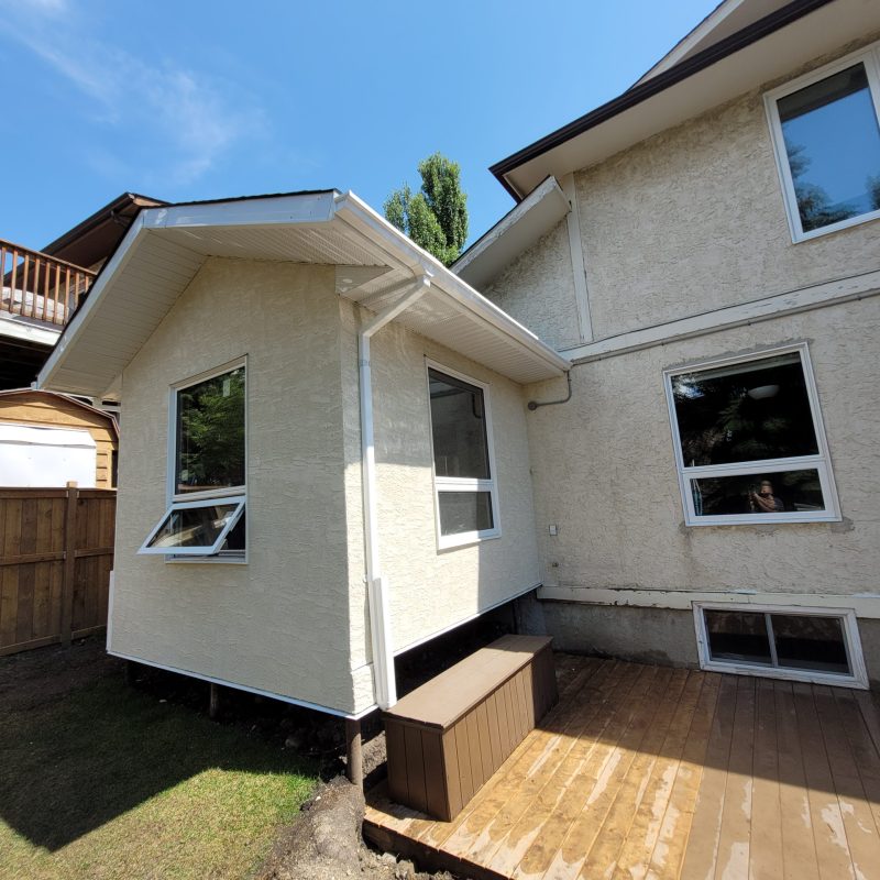 Exterior view of a beige stucco house featuring a small room addition with open awning windows, next to a wooden deck with a storage bench.