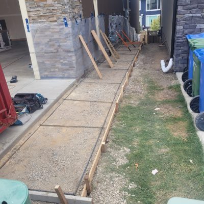 Construction site view of a walkway preparation next to a garage, featuring a rebar grid laid within wooden forms, plastic sheeting protecting a stone veneer column, and recycling bins stored on the side.